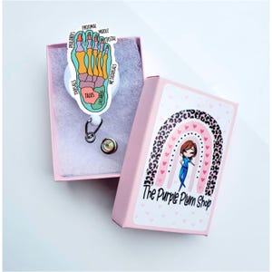 May include: A pink box with a rainbow and leopard print design. The box has the text "The Purple Plum Shop" on it. The box contains a white retractable badge reel with a colourful anatomical foot illustration.