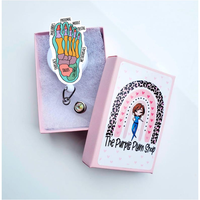 May include: A pink box with a rainbow and leopard print design. The box has the text "The Purple Plum Shop" on it. The box contains a white retractable badge reel with a colourful anatomical foot illustration.