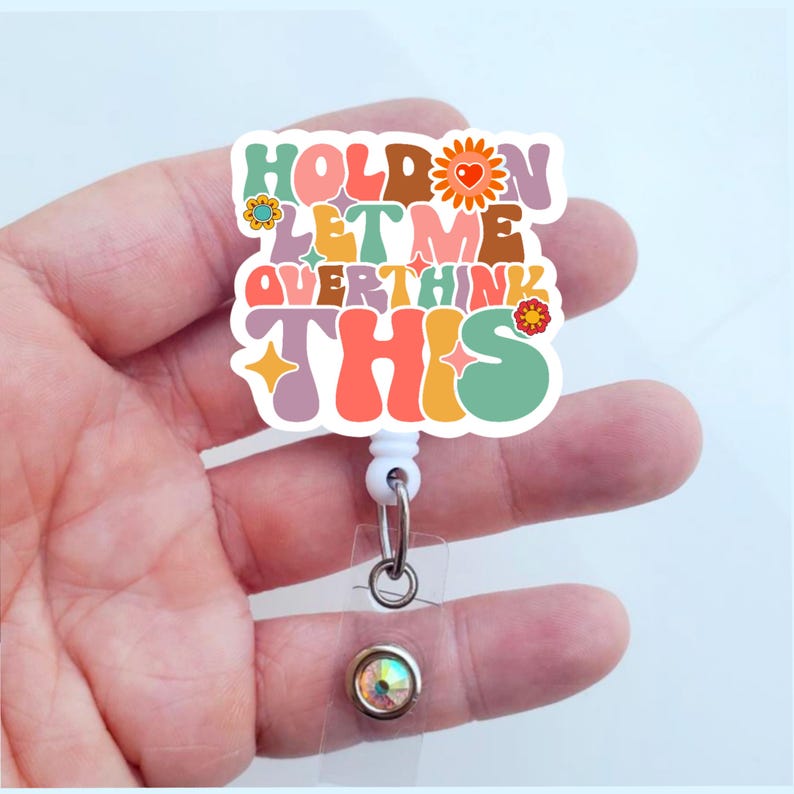 May include: A colorful badge reel with the text "Hold on let me overthink this" in a retro font. The reel has a white retractable cord and a clear plastic badge holder with a sparkly rhinestone.