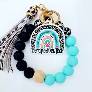 May include: A turquoise and black beaded keychain with a gold key ring and a charm featuring a rainbow with a paw print and the text "Let all you do be done in love 1 Corinthians 16:14" and "Certified Vet Tech".