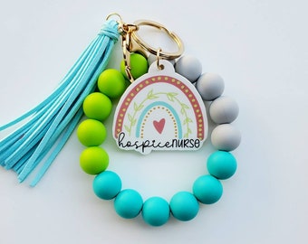May include: A light blue tassel keychain with a rainbow charm and the words "hospice nurse". The charm is surrounded by green, turquoise, and grey silicone beads.