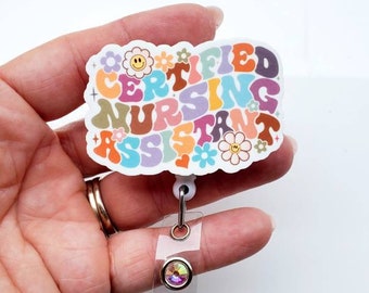 May include: A colorful badge reel with the words "CERTIFIED NURSING ASSISTANT" in a retro font. The badge has a clear plastic strap and a decorative, iridescent button. The design includes flowers and a smiling face.