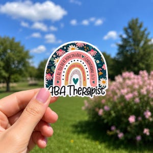 May include: A semi-circular sticker featuring a dark blue background with colorful flowers, a central rainbow design, and the text "LET ALL YOU DO BE DONE IN LOVE 1 CORINTHIANS 16:14" and "ABA Therapist" in black script lettering.