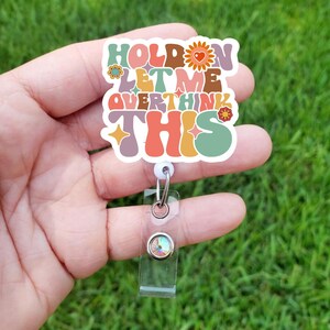 May include: A colorful badge holder with the text "Hold on let me overthink this" in a retro font. The badge holder has a clear plastic strap and a sparkly rhinestone.