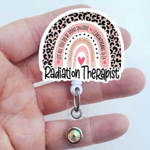 May include: A badge reel with a rainbow design and the words "Radiation Therapist." The rainbow features a leopard print border, pink and brown arches, and the text "Let all you do be done in love 1 Corinthians 16:14."