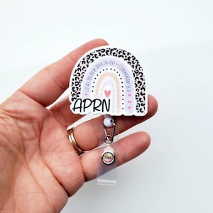 May include: A white and black leopard print rainbow with the text "Let all you do be done in love" and "I Corinthians 16:14" around the rainbow. The rainbow has a pink heart in the centre and a white background. The text "APRN" is below the rainbow. The rainbow is attached to a clear retractable badge holder with a rainbow rhinestone.