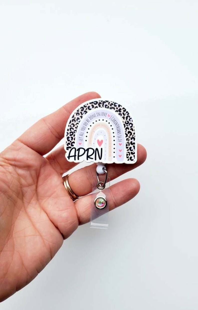 May include: A white and black leopard print rainbow with the text "Let all you do be done in love" and "I Corinthians 16:14" around the rainbow. The rainbow has a pink heart in the centre and a white background. The text "APRN" is below the rainbow. The rainbow is attached to a clear retractable badge holder with a rainbow rhinestone.