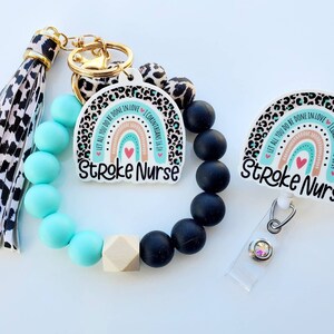 May include: A set of accessories for a nurse, including a keychain with a tassel, a beaded bracelet, and a badge reel. The items feature a rainbow design with the text "Stroke Nurse" and a Bible verse. The bracelet has turquoise and black beads.