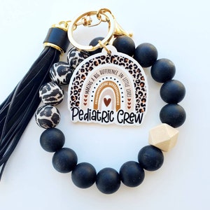 May include: A black beaded bracelet with a wooden bead and a keychain charm. The charm features a rainbow with the text "Making a big difference in little lives" and "Pediatric Crew".