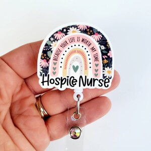 May include: A white and pink rainbow with a floral pattern and the text "Because your life is worth my time" and "Hospice Nurse". The rainbow is attached to a clear plastic badge holder with a sparkly rhinestone.