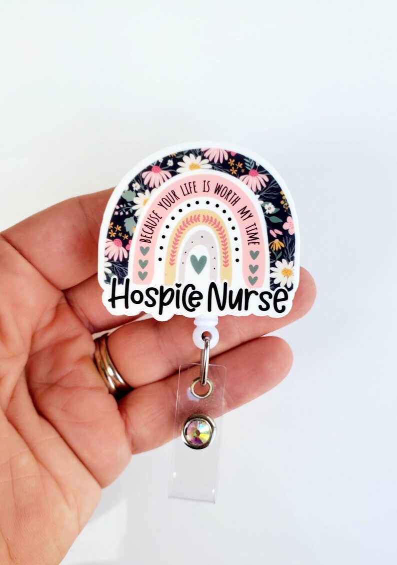 May include: A white and pink rainbow with a floral pattern and the text "Because your life is worth my time" and "Hospice Nurse". The rainbow is attached to a clear plastic badge holder with a sparkly rhinestone.