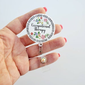 May include: White round badge holder with a floral design and the text "Occupational Therapy" surrounded by positive attributes. The badge holder is attached to a clear retractable badge reel with a sparkly rhinestone.