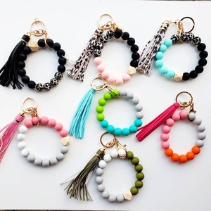 May include: Assortment of colorful beaded keychains with tassels. Each keychain features a circle of beads with a wooden accent and a gold key ring.