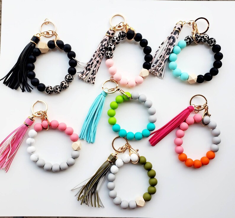 May include: Assortment of colorful beaded keychains with tassels. Each keychain features a circle of beads with a wooden accent and a gold key ring.
