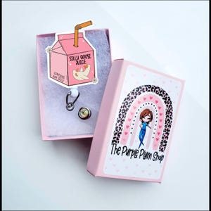May include: A pink box with a cartoon goose on the lid. The box says "Silly Goose Juice" and "Contains real silly". The box is open and shows a silver retractable badge reel with a clear, round charm. The box is sitting on a white surface. The box is decorated with a rainbow and leopard print. The text "The Purple Plum Shop" is on the box.