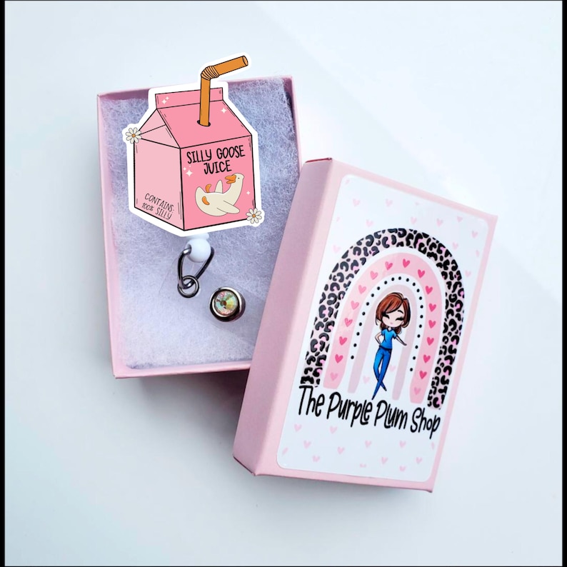 May include: A pink box with a cartoon goose on the lid. The box says "Silly Goose Juice" and "Contains real silly". The box is open and shows a silver retractable badge reel with a clear, round charm. The box is sitting on a white surface. The box is decorated with a rainbow and leopard print. The text "The Purple Plum Shop" is on the box.