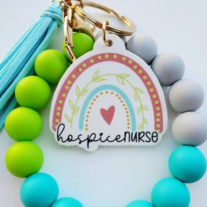 May include: A colorful rainbow charm with the text "hospice nurse" attached to a beaded bracelet with turquoise, green, and gray beads. The bracelet has a gold key ring attached.