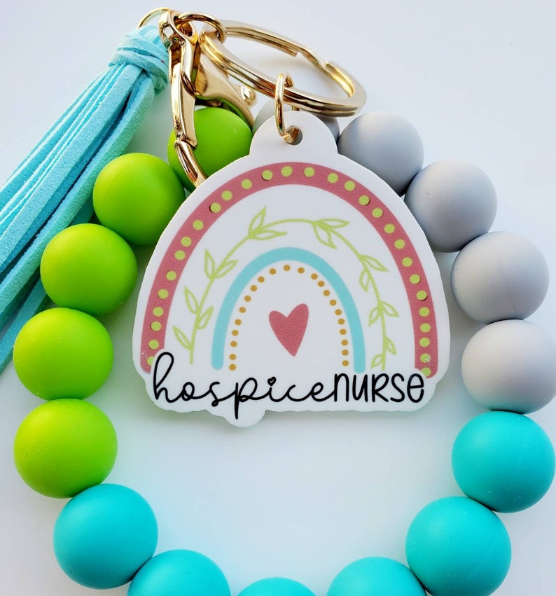 May include: A colorful rainbow charm with the text "hospice nurse" attached to a beaded bracelet with turquoise, green, and gray beads. The bracelet has a gold key ring attached.