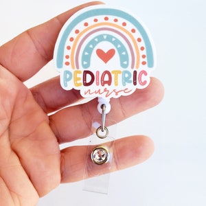 May include: A white badge reel with a rainbow design and the text "PEDIATRIC nurse". The rainbow has a red heart in the center. The badge reel has a clear plastic strap and a clear plastic clip with a gold-colored ring and a clear rhinestone.