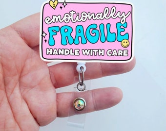 May include: Pink badge holder with a white retractable reel and a sparkly rhinestone. The badge holder has a pink background with the text "emotionally FRAGILE HANDLE WITH CARE" in white and blue lettering.