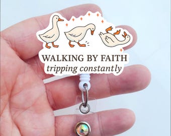 May include: A white badge reel with a cartoon goose design. The design shows three geese in different poses, with the text "WALKING BY FAITH tripping constantly". The reel has a clear plastic strap and a decorative iridescent button.
