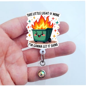 Op de afbeelding: Een witte intrekbare badgehouder met een groene cartoon-vuilnisbak met vlammen erop. De vuilnisbak heeft een vrolijk gezicht en de tekst "This little light of mine I'm gonna let it shine".