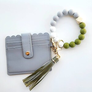 May include: A gray leather card holder with a scalloped edge and a green tassel keychain. The keychain has a beaded bracelet with gray, white, and green beads.