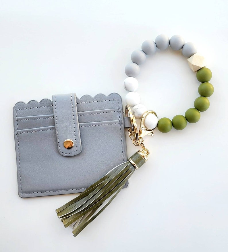 May include: A gray leather card holder with a scalloped edge and a green tassel keychain. The keychain has a beaded bracelet with gray, white, and green beads.