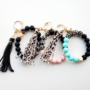 May include: Three beaded keychains with tassels. One keychain has black beads, one has black and white leopard print beads, and one has pink and turquoise beads.