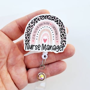 May include: A white and pink rainbow with a leopard print border and the text "Let all you do be done in love 1 Corinthians 16:14" and "Nurse Manager". The rainbow has a pink heart in the center. The badge holder has a clear plastic strap and a silver retractable reel with a clear crystal.