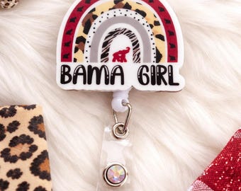 May include: A white badge reel with a rainbow design and the text "BAMA GIRL". The rainbow features red, leopard print, and gray patterns. The reel has a clear plastic strap and a decorative gem.