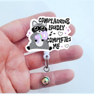 May include: A white retractable badge holder with a clear plastic charm featuring a cartoon raccoon holding a megaphone with the text "Complaining Loudly Completes Me" in black text with pink hearts and stars.