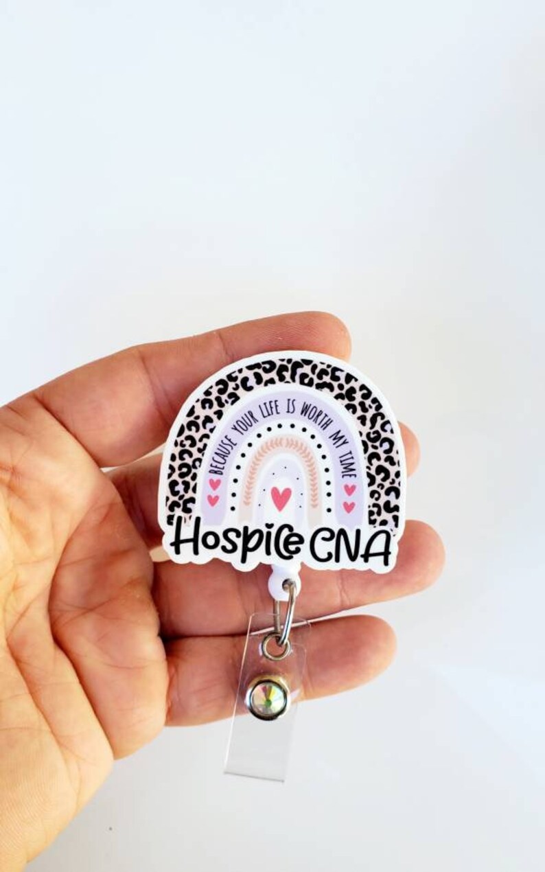 May include: A white and black leopard print rainbow with the text "Because your life is worth my time" and three pink hearts. The rainbow is attached to a white retractable badge holder with a clear plastic strap and a silver clip. The text "Hospice CNA" is printed below the rainbow.