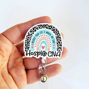 May include: A white and blue rainbow with a leopard print background. The rainbow has a pink and peach center. The text "Because your life is worth my time" is written on the rainbow. The text "Hospice CNA" is written below the rainbow. A clear badge holder with a silver clip and a clear rhinestone is attached to the bottom of the rainbow.