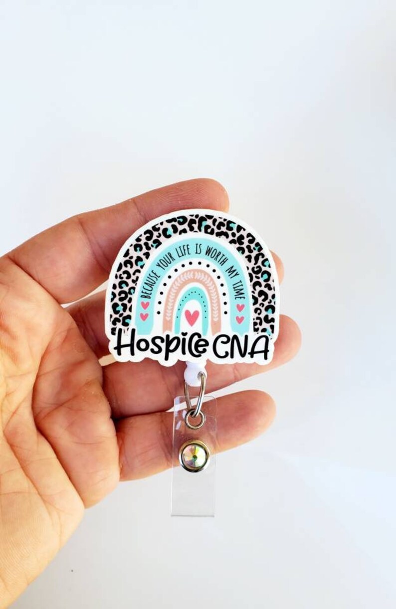May include: A white and blue rainbow with a leopard print background. The rainbow has a pink and peach center. The text "Because your life is worth my time" is written on the rainbow. The text "Hospice CNA" is written below the rainbow. A clear badge holder with a silver clip and a clear rhinestone is attached to the bottom of the rainbow.