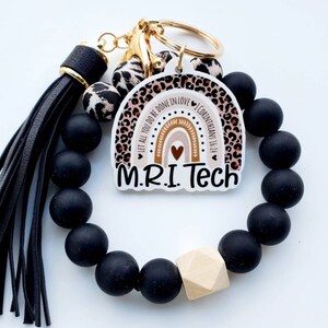 May include: A black beaded bracelet with a wooden bead and a keychain charm. The charm features a rainbow with the text "Let all you do be done in love 1 Corinthians 16:14" and "MRI Tech".