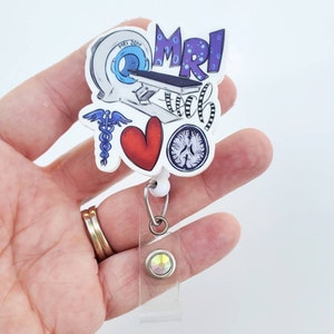 May include: A white plastic badge holder with a colorful design featuring an MRI machine, a heart, a brain, a caduceus, and the text "MRI Tech". The badge holder has a clear plastic strap and a silver clip with a clear rhinestone.
