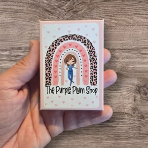 May include: A pink and white box with a rainbow design featuring a cartoon nurse and the text "The Purple Plum Shop".