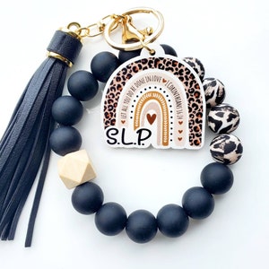 May include: Black beaded bracelet with a wooden bead and a keychain charm featuring a rainbow with a leopard print background. The charm also includes the text "Let all you do be done in love 1 Corinthians 16:14" and "SLP".
