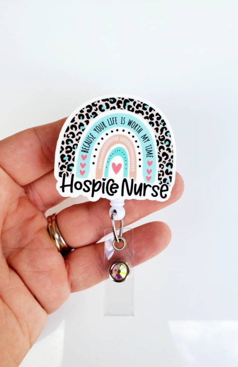 May include: A white and blue rainbow with a leopard print border and the text "Because your life is worth my time" and "Hospice Nurse". The rainbow has a pink and peach center and pink hearts. The badge holder has a clear plastic strap and a clear plastic badge holder with a sparkly gem.