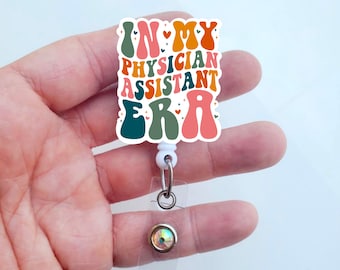 Puede incluir: Un portagafete retráctil blanco con una pegatina colorida de estilo retro que dice "In My Physician Assistant Era". La pegatina tiene un esquema de color verde, naranja, rosa y amarillo con letras blancas. El portagafete tiene una cubierta de plástico transparente y un strass brillante.