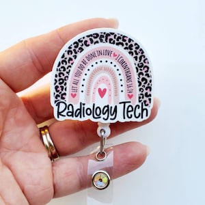 May include: A white and pink rainbow with a leopard print border and the text "Let all you do be done in love 1 Corinthians 16:14" and "Radiology Tech". The rainbow has a pink heart in the center. The badge holder has a clear plastic strap and a silver clip with a crystal.