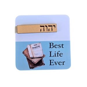 May include: A wooden bookmark with the Hebrew words "Best Life Ever" and a depiction of a book with the words "Holy Scriptures" on the cover. The bookmark is on a blue background.