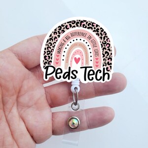 May include: A pink and brown rainbow with leopard print accents and the text "Making a Big Difference in Little Lives" and "Peds Tech". The rainbow has a white background and is attached to a clear retractable badge holder with a sparkly rhinestone.
