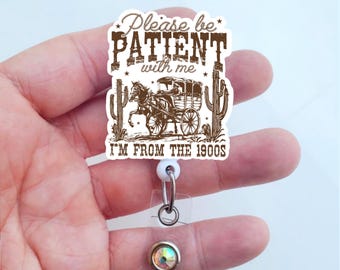 May include: White plastic badge holder with a brown and white graphic that says "Please be PATIENT with me I'M FROM THE 1900s". The graphic features a horse-drawn carriage and cacti.