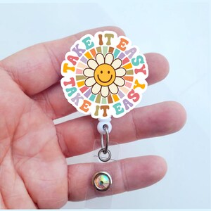 May include: A white retractable badge holder with a colorful flower design. The flower has a smiling yellow face and the text "Take it easy" is written around the flower.
