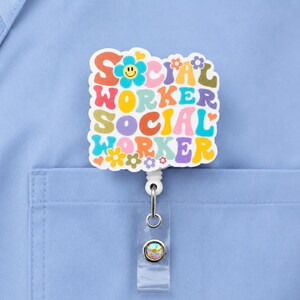 May include: A colorful badge reel with the words "SOCIAL WORKER" in a retro font, featuring flowers and a smiling flower. The badge is attached to a retractable reel with a clear plastic strap and a decorative gem.
