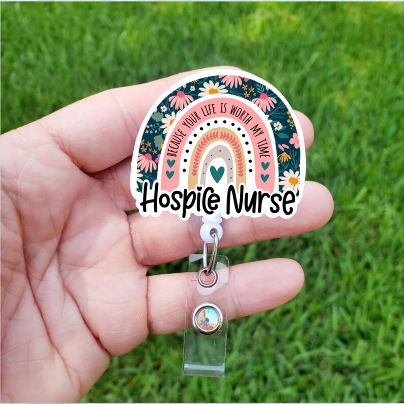 May include: A pink and green floral rainbow with the text "Because your life is worth my time" and "Hospice Nurse" on a white background. The rainbow is attached to a clear plastic retractable badge holder with a silver clip.