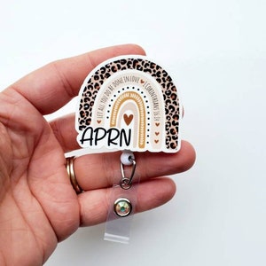 May include: A brown and tan leopard print rainbow with the text "Let all you do be done in love" and "1 Corinthians 16:14" around the rainbow. The text "APRN" is below the rainbow. The rainbow is attached to a clear retractable badge holder with a sparkly blue gem.