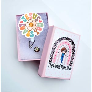 May include: A pink box with a rainbow and leopard print design. The box says "The Purple Plum Shop". The box contains a white retractable badge reel with a colorful flower and the words "Jesus Loves You".